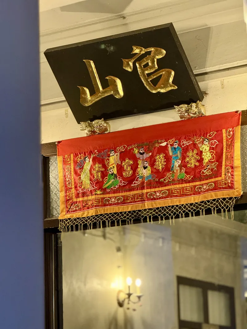 ป้ายไม้กวนซ้าน อักษรจีนทองบนพื้นดำ เหนือประตูบ้านอาจ้อ พร้อมผ้าแดงปักลาย